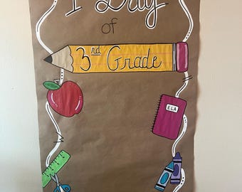 Puede incluir: Un letrero de papel marrón con las palabras "1st Day of 3rd Grade" en negro y amarillo. El letrero está decorado con un lápiz, una manzana, un cuaderno, crayones y otras ilustraciones escolares.