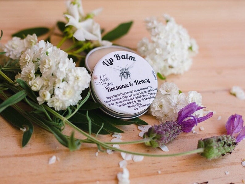 Ninas Bees beeswax lip balm is in a compact aluminium tin with a paper label. it rests among flowers to symbolise its nature: pure botanical ingredients.