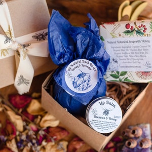 Spa gift for her. the gift contains bath bomb wrapped in blue tissue paper with a  Bee label. there is a honey lip balm and honey soap bar in the box. the soap bar is wrapped in a paper with floral pattern. all packaging is environmentally friendly