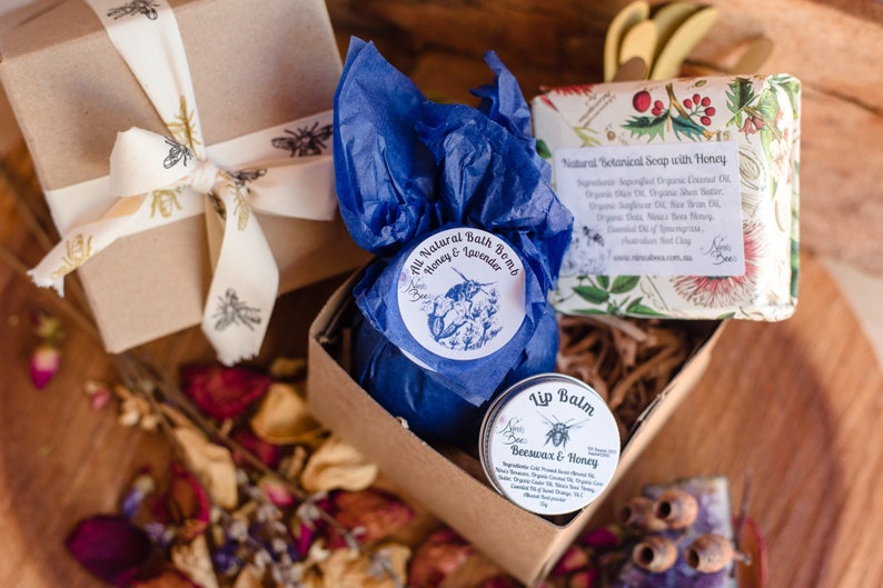Spa gift for her. the gift contains bath bomb wrapped in blue tissue paper with a  Bee label. there is a honey lip balm and honey soap bar in the box. the soap bar is wrapped in a paper with floral pattern. all packaging is environmentally friendly
