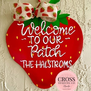 May include: Red and green strawberry-shaped wooden door hanger with a white bow and the text "Welcome to our Patch The Halstroms".