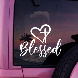May include: White vinyl decal featuring the word "Blessed" in a cursive font, with a heart and cross design above. The decal is displayed on a pink vehicle window, with a beach scene visible in the background.