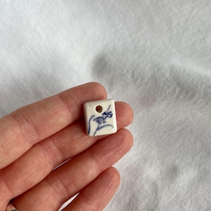 Può includere: Un piccolo ciondolo quadrato in ceramica bianca con un motivo floreale blu e un foro nella parte superiore. Il ciondolo è tenuto in mano su uno sfondo bianco. Il motivo floreale è delicato e dettagliato.