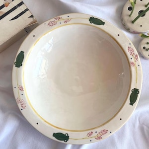 Puede incluir: Un cuenco de cerámica color crema con un borde decorativo. El borde presenta ilustraciones de jarrones verdes, acentos florales rosas y puntos negros. Una fina línea dorada bordea el borde. Ideal para servir o decorar.