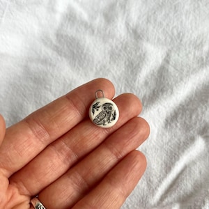 May include: A small, circular pendant featuring a black and white owl design. The pendant is white with a small loop at the top. The owl is depicted on a branch with leaves. The pendant is held in a hand.
