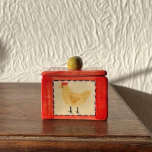 Peut inclure: Petite boîte rectangulaire en céramique avec couvercle. Rouge, avec un carré représentant une poule jaune sur fond blanc. Le couvercle a un petit bouton rond vert olive. Posée sur une surface en bois.