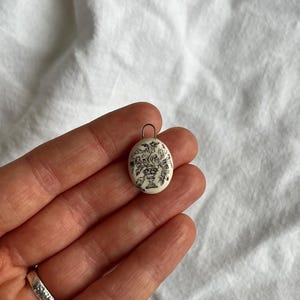 Può includere: Ciondolo ovale con un design floreale in bianco e nero. Il ciondolo presenta un'illustrazione dettagliata di fiori in un vaso. Il ciondolo è tenuto in una mano su uno sfondo bianco. Un piccolo anello di metallo è attaccato in alto.