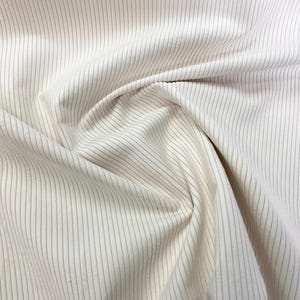 K&ouml;nnte beinhalten: Nahaufnahme eines cremefarbenen Stoffes mit d&uuml;nnen, vertikalen grauen Streifen. Der Stoff ist drapiert und gefaltet, wodurch Schatten und Textur entstehen. Die Streifen sind gleichm&auml;&szlig;ig verteilt und verlaufen &uuml;ber die gesamte L&auml;nge des Materials.