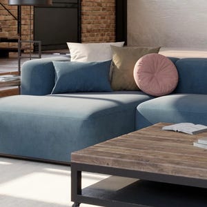 K&ouml;nnte beinhalten: Ein modernes Wohnzimmer mit einem gro&szlig;en, blauen Ecksofa und mehreren dekorativen Kissen in neutralen T&ouml;nen. Vor dem Sofa steht ein Couchtisch aus Holz mit schwarzem Metallrahmen. Der Raum hat eine Backsteinmauer und eine gro&szlig;e Lampe.