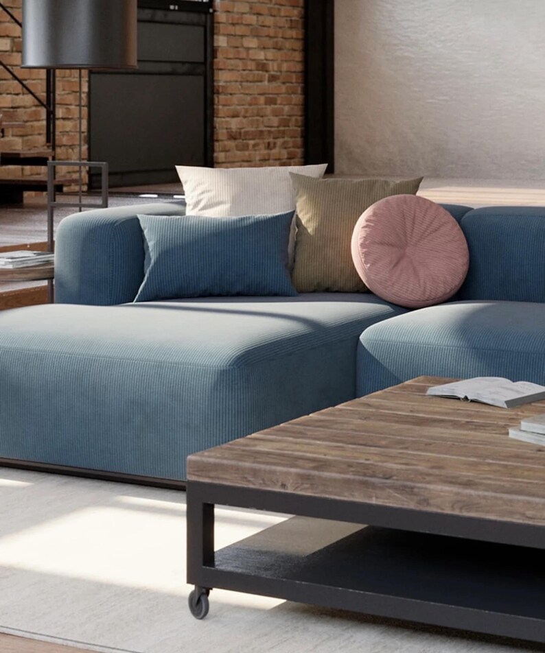 K&ouml;nnte beinhalten: Ein modernes Wohnzimmer mit einem gro&szlig;en, blauen Ecksofa und mehreren dekorativen Kissen in neutralen T&ouml;nen. Vor dem Sofa steht ein Couchtisch aus Holz mit schwarzem Metallrahmen. Der Raum hat eine Backsteinmauer und eine gro&szlig;e Lampe.