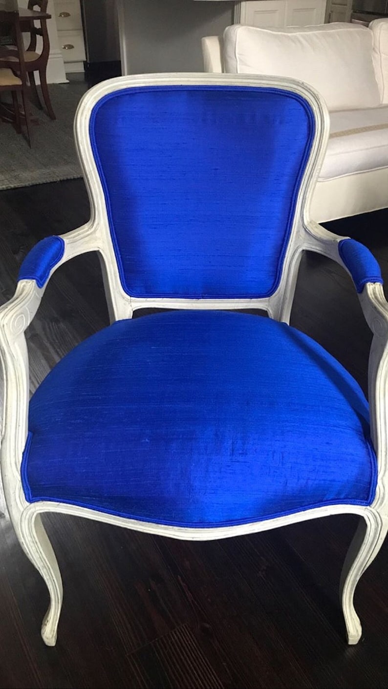 Puede incluir: Un sill&oacute;n de madera blanca con un asiento y respaldo de terciopelo azul. El sill&oacute;n tiene un respaldo y brazos curvos, y las patas est&aacute;n talladas en un estilo tradicional.