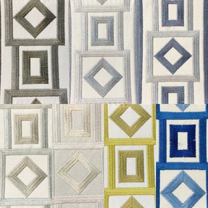May include: A close-up of a fabric with a geometric pattern of squares and diamonds in shades of gray, blue, and green. The fabric has a white background and the pattern is embroidered.