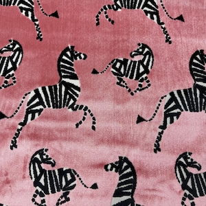 May include: Pink fabric with a repeating pattern of black and white zebras in various poses. The zebras are stylized with bold stripes, and the fabric has a soft, velvety texture. This design is suitable for home decor or fashion.