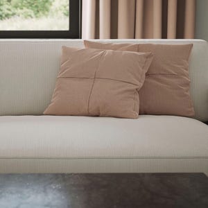 K&ouml;nnte beinhalten: Zwei quadratische, altrosa Kissen mit vertikaler Rippenstruktur und einer mittigen Kreuznaht, die auf einem hellbeigen, fein gestreiften Sofa mit einem dezenten, gleichm&auml;&szlig;igen Stoffmuster liegen.
