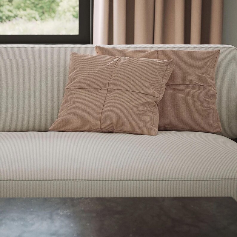 K&ouml;nnte beinhalten: Zwei quadratische, altrosa Kissen mit vertikaler Rippenstruktur und einer mittigen Kreuznaht, die auf einem hellbeigen, fein gestreiften Sofa mit einem dezenten, gleichm&auml;&szlig;igen Stoffmuster liegen.
