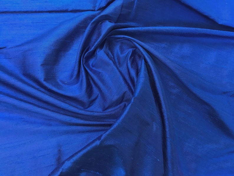 Puede incluir: Primer plano de una tela lisa de color azul real con un brillo sedoso. La tela est&aacute; drapeada y doblada, creando l&iacute;neas suaves y fluidas.