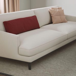K&ouml;nnte beinhalten: Ein cremefarbenes Sofa mit einem dunkelbraunen Rahmen und zwei Wurfkissen. Ein rechteckiges, weinrotes Kissen liegt auf der linken Seite. Das Sofa steht auf einem gemusterten Teppich und schafft einen neutralen und einladenden Wohnraum.