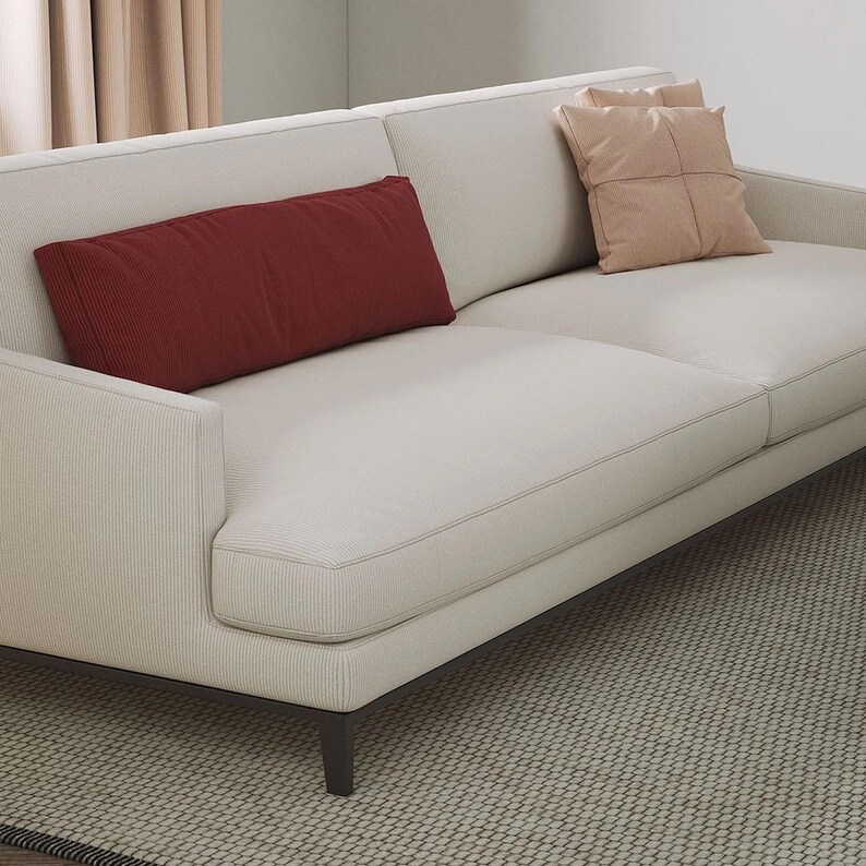 K&ouml;nnte beinhalten: Ein cremefarbenes Sofa mit einem dunkelbraunen Rahmen und zwei Wurfkissen. Ein rechteckiges, weinrotes Kissen liegt auf der linken Seite. Das Sofa steht auf einem gemusterten Teppich und schafft einen neutralen und einladenden Wohnraum.