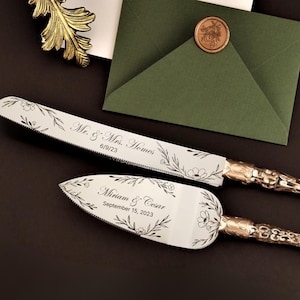 Puede incluir: Un juego de cuchillo y servidor de pastel plateado con grabado floral y mangos dorados. El cuchillo está inscrito con "Mr. & Mrs. Homes 6/9/23" y el servidor con "Miriam & Cesar September 15, 2023". Un sobre verde y un sello de cera en el fondo.
