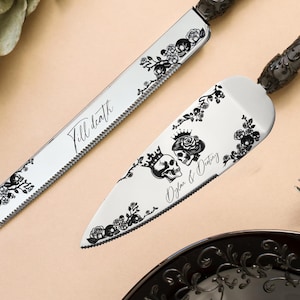 Op de afbeelding: Een bruidstaartmes en serveerset met zwarte bloemen- en schedelontwerpen. Op het mes staat "Till death" en op de serveerschaal "Officiant & Destiny". Twee vorken en een zwart bord zijn ook zichtbaar.