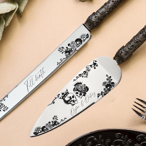 Op de afbeelding: Een bruidstaartmes en -serveerset met zwarte bloemen- en schedelontwerpen. Op het mes staat "Till death" en op de serveerder "Of life & Destiny". De set bevat twee vorken en een zwart bord.