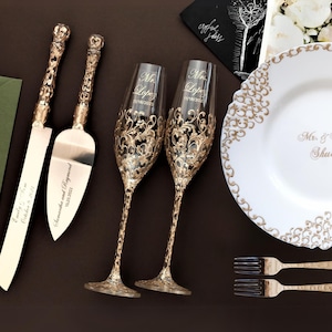 Könnte beinhalten: Ein weißer Teller mit goldener Dekorrand und dem Text "Mr. & Mrs. Schultz" in goldenen Lettern. Zwei goldgeränderte Champagnerflöten, ein goldgriffiges Kuchenschwert und ein -server sowie zwei goldene Gabeln sind auf einer braunen Oberfläche angeordnet.