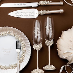May include: Wedding table setting with a cake knife and server, two champagne flutes, a ring pillow, and a plate with forks. The items are decorated with pearls and sit on a brown surface. The knife and server have text.