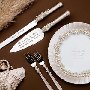 Könnte beinhalten: Ein Hochzeitstorten-Servierset mit Perlenakzenten, einschließlich Messer und Servierer, sowie zwei Gabeln und einem dekorativen Teller. Das Set befindet sich auf einer braunen Oberfläche mit Text.