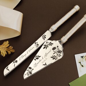 Puede incluir: Un juego de cuchillo y servidor para pastel de bodas con mangos de marfil y grabado floral. El cuchillo y el servidor están decorados con diseños florales negros y los nombres "Macy & Steven" y "Brandon & Mariah 10.04.25".