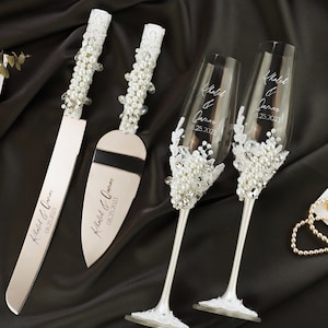 May include: A wedding cake knife and server set with two champagne flutes. The knife and server have pearl and crystal accents on the handles. The flutes are decorated with pearls and lace. Each item is engraved with a name and date.