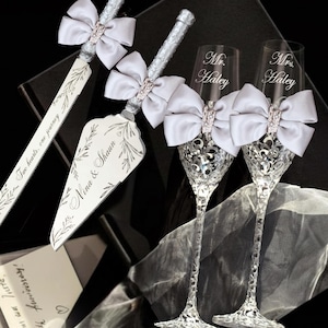 Könnte beinhalten: Ein Hochzeitstortenmesser- und Servier-Set mit silbernen Griffen und weißen Schleifen, die mit Strasssteinen verziert sind. Zwei Sektflöten mit Kristallfüßen und passenden Schleifen sind ebenfalls vorhanden. Das Set wird in einer schwarzen Box präsentiert.