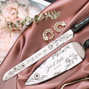 May include: A set of two silver cake knives with black floral handles. The knives are engraved with the text "To Be With You" and "Jake & Nikki 10-21-23" along with floral designs.