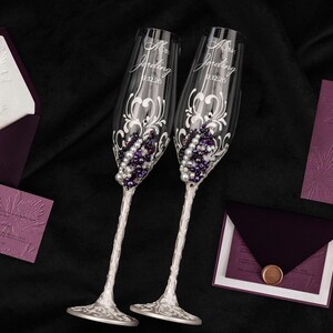 May include: Two clear glass champagne flutes featuring white and purple bead embellishments, silver stems, and etched text reading Mr. Jording and Mrs. Jording with the date 9.12.20, accompanied by dark purple and white wedding stationery sets.