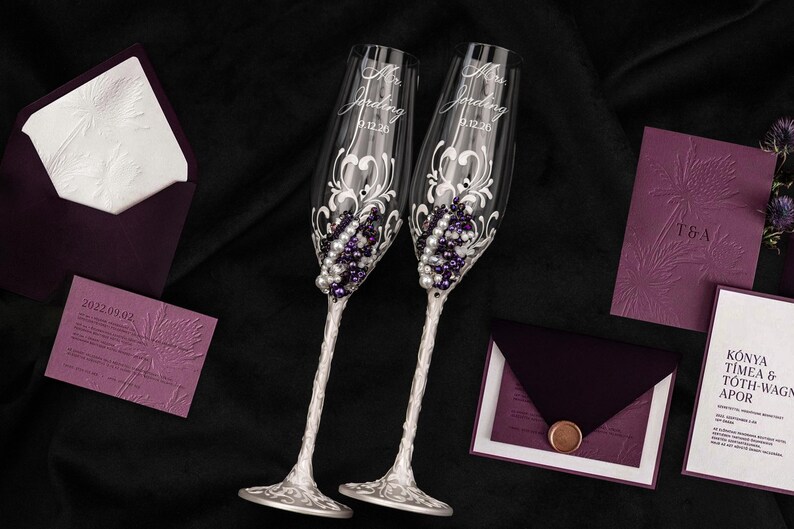 May include: Two clear glass champagne flutes featuring white and purple bead embellishments, silver stems, and etched text reading Mr. Jording and Mrs. Jording with the date 9.12.20, accompanied by dark purple and white wedding stationery sets.
