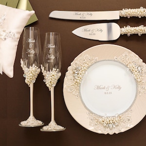 Puede incluir: Un conjunto de boda que incluye un coj&iacute;n para anillos color crema, dos copas de champ&aacute;n, un cuchillo y pala para pastel, un plato y dos tenedores. Cada pieza est&aacute; decorada con perlas blancas, cristales y encaje, con el texto Mark & Kelly 9.2.23.