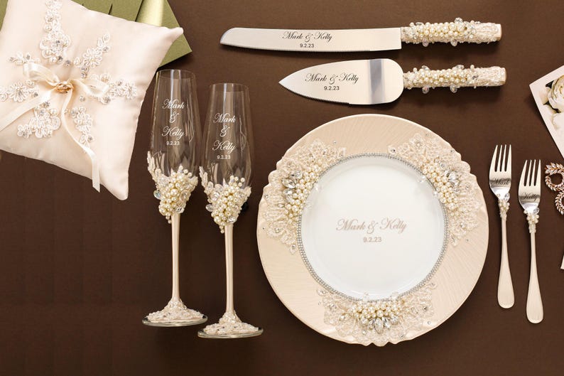 Puede incluir: Un conjunto de boda que incluye un coj&iacute;n para anillos color crema, dos copas de champ&aacute;n, un cuchillo y pala para pastel, un plato y dos tenedores. Cada pieza est&aacute; decorada con perlas blancas, cristales y encaje, con el texto Mark & Kelly 9.2.23.