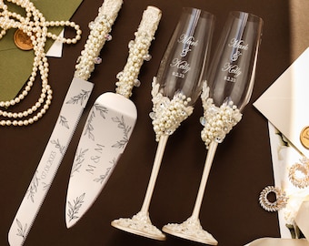 May include: A wedding cake cutting set with a silver knife and server with a floral design and the initials M & M and the date 07.01.2023. Two champagne flutes with pearl embellishments and the names Mark & Kelly and the date 8.2.23.