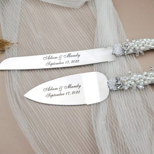 May include: A silver cake cutting set with pearl and lace handles. The blades are engraved with "Adam & Mandy September 17, 2022".