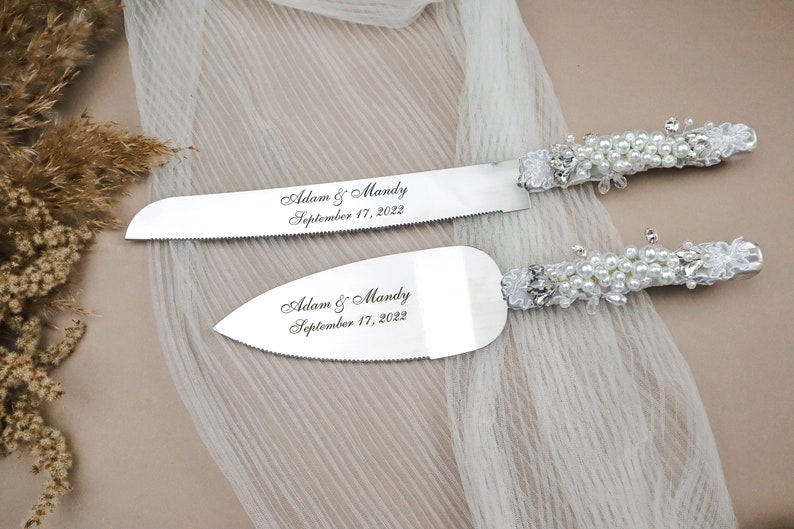May include: A silver cake cutting set with pearl and lace handles. The blades are engraved with "Adam & Mandy September 17, 2022".