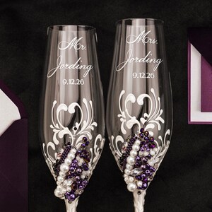 May include: Two clear glass champagne flutes featuring white swirling filigree patterns, clusters of white pearls, and deep purple beads. Each glass is etched with the text Mr. Jording or Mrs. Jording and the date 9.12.26 in a cursive script font.