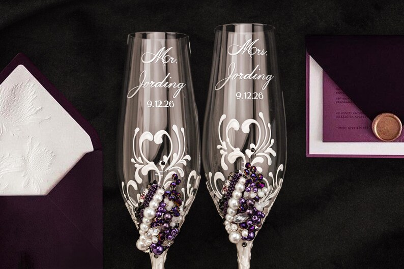 May include: Two clear glass champagne flutes featuring white swirling filigree patterns, clusters of white pearls, and deep purple beads. Each glass is etched with the text Mr. Jording or Mrs. Jording and the date 9.12.26 in a cursive script font.