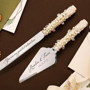 May include: A silver cake knife and server set with handles embellished with white pearls, clear crystals, and lace. The knife blade is engraved with the text You are my greatest adventure, and the server features the names Rosalia & Trevor 09.23.2023.