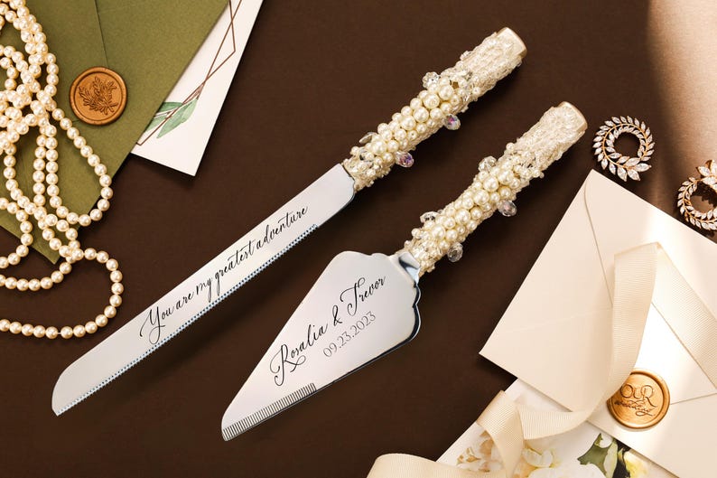 May include: A silver cake knife and server set with handles embellished with white pearls, clear crystals, and lace. The knife blade is engraved with the text You are my greatest adventure, and the server features the names Rosalia & Trevor 09.23.2023.