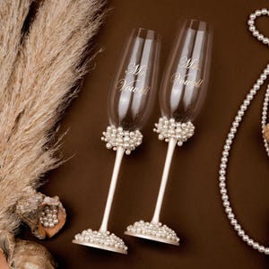 Puede incluir: Dos copas de champ&aacute;n con detalles de perlas y las palabras "Mr. Vow" y "Mrs. Vow" grabadas en el cristal. Las copas tienen tallos blancos y bases adornadas con perlas. La imagen incluye conchas marinas, perlas y plantas secas sobre un fondo marr&oacute;n.