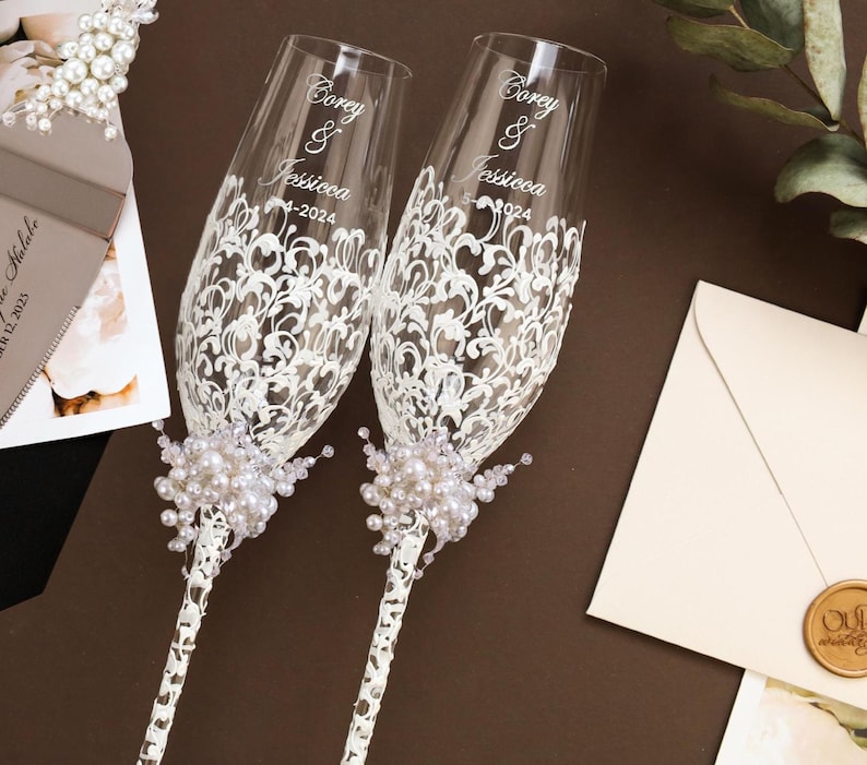 May include: Two champagne flutes with white lace design and pearl accents. The flutes are personalized with the names "Corey & Jessica" and the date "5-4-2024". A wedding cake server and envelope are also visible.