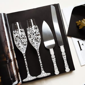 Könnte beinhalten: Elegante Sektgläser und Servierutensilien mit aufwendigen weißen Details. Das Set umfasst zwei Gläser, einen Tortenheber und ein Messer, alle auf einer schwarzen Oberfläche angeordnet. Ein schwarzer Umschlag mit goldenem Siegel und ein goldenes Blatt sind ebenfalls vorhanden.