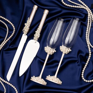 Puede incluir: Un juego de cuchillo y servidor para pastel de bodas con dos copas de champ&aacute;n, todos adornados con perlas de imitaci&oacute;n. Los mangos del cuchillo y el servidor est&aacute;n tachonados de perlas, al igual que las bases de las copas. Sobre sat&eacute;n azul oscuro.