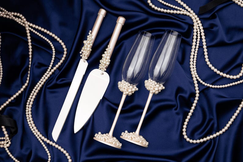 Puede incluir: Un juego de cuchillo y servidor para pastel de bodas con dos copas de champ&aacute;n, todos adornados con perlas de imitaci&oacute;n. Los mangos del cuchillo y el servidor est&aacute;n tachonados de perlas, al igual que las bases de las copas. Sobre sat&eacute;n azul oscuro.