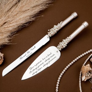 May include: A silver wedding cake knife and server set with pearl accents. The knife and server have engraved text, and the set is displayed with pearls and seashells on a brown surface.