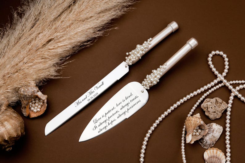 May include: A silver wedding cake knife and server set with pearl accents. The knife and server have engraved text, and the set is displayed with pearls and seashells on a brown surface.